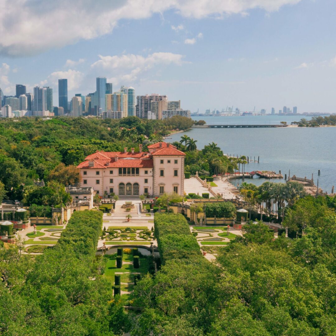 Aerial view of the historic villa at Vizcaya with gardens by the waterfront, set against a city skyline and blue sky, perfect for celebrating Labor Day.