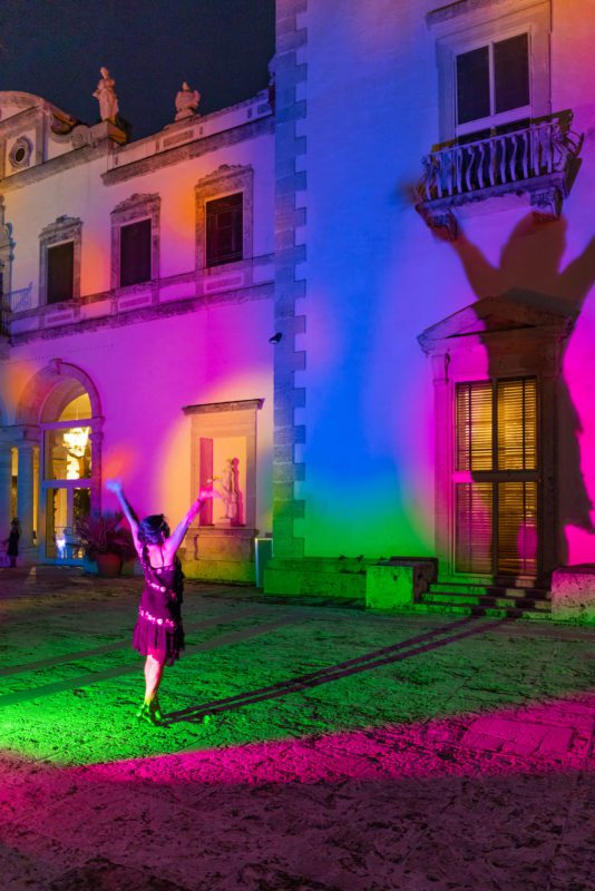 Person standing with arms up with shadow showing on Vizcaya's Main House lighted up in red, blue, yellow and green.