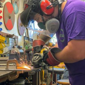 Man in a protective mask and helmet uses a saw to work a piece of metal in a blacksmith shop