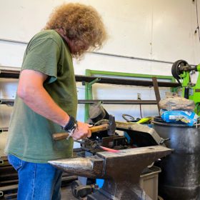 Blacksmith shop featuring a man with with curly brown hair working a piece of hot metal on an anvil