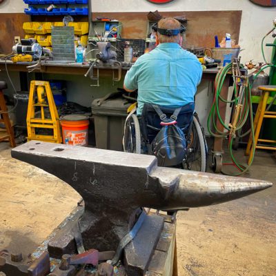 Man in a wheelchair with head gear behind a large anvil