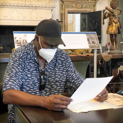 A Black man in a hat and a mask reads over historic documents
