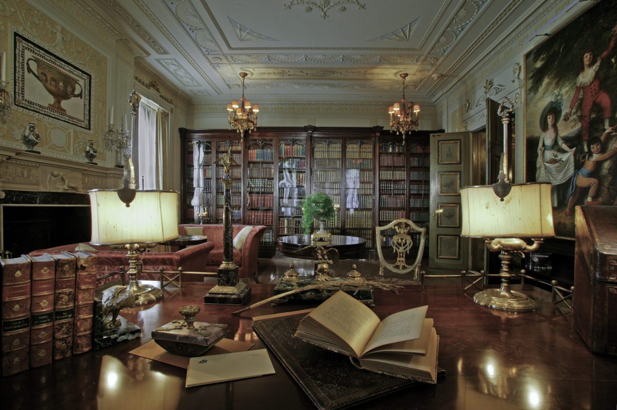 A library featuring a desk with different books and papers. The room also features a fireplace, decorative paintings and statues and a wall of books