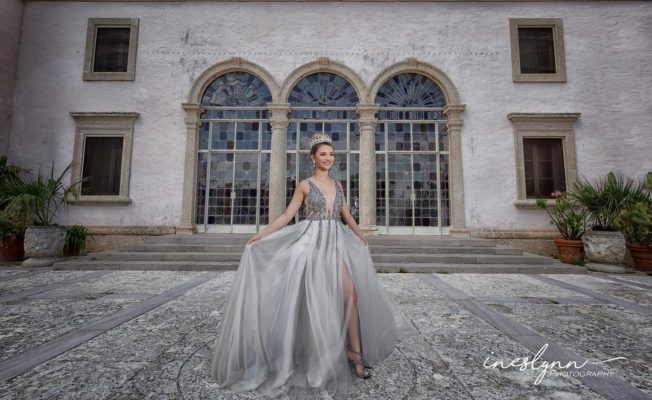 Glowing young girl wearing a tiara and a sparkling silver dress poses for quince photos outside the doors of the Enclosed Loggia at Vizcaya. Photo by Ineslynn Photography
