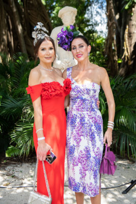 A lady in a red dress and a women in a purple dress and matching hat.