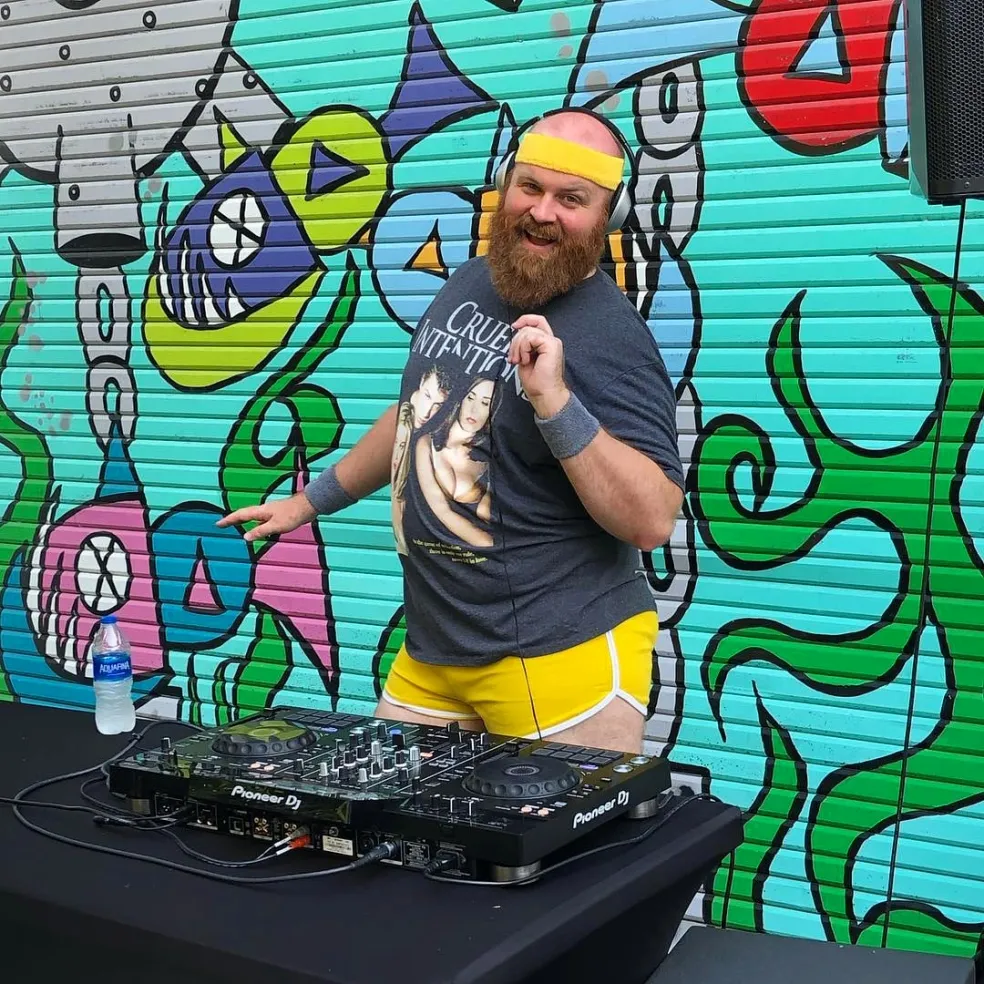 A DJ stands in yellow hotpants in front of an audio mixerboard. Behind him is a vibrant and colorful street art mural