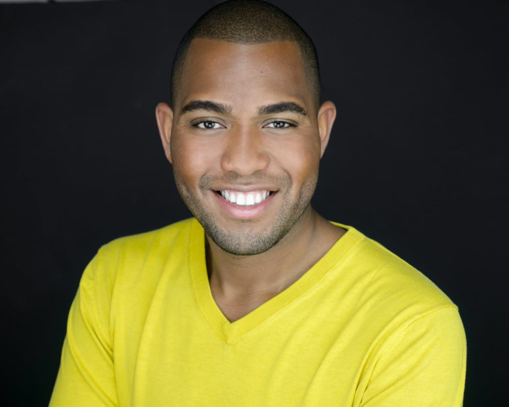A Man of color wearing a bright yellow shirt in front of a black background