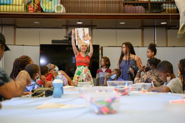 Artist holding up image with colorful patterns to group including several families. Art tools are on a table. Workshop taking place in Vizcaya's Historic Garage.