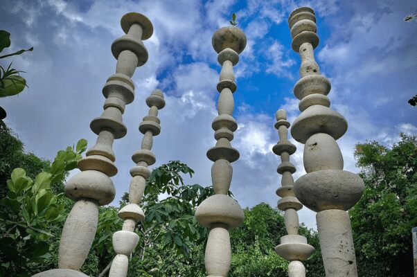 Tall stone pillars with intricate carvings rise against a backdrop of cloudy sky and surrounding greenery.