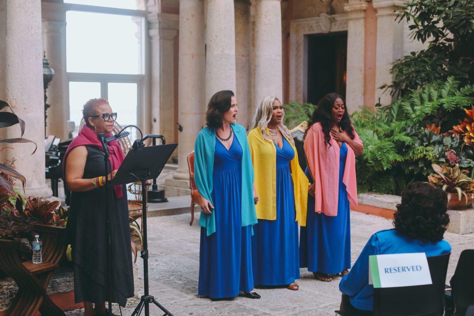 One woman at a microphone doing dramatic reading. Three women in blue dresses and three different colored scarfs singing. All in the Courtyard of Vizcaya Museum and Gardens