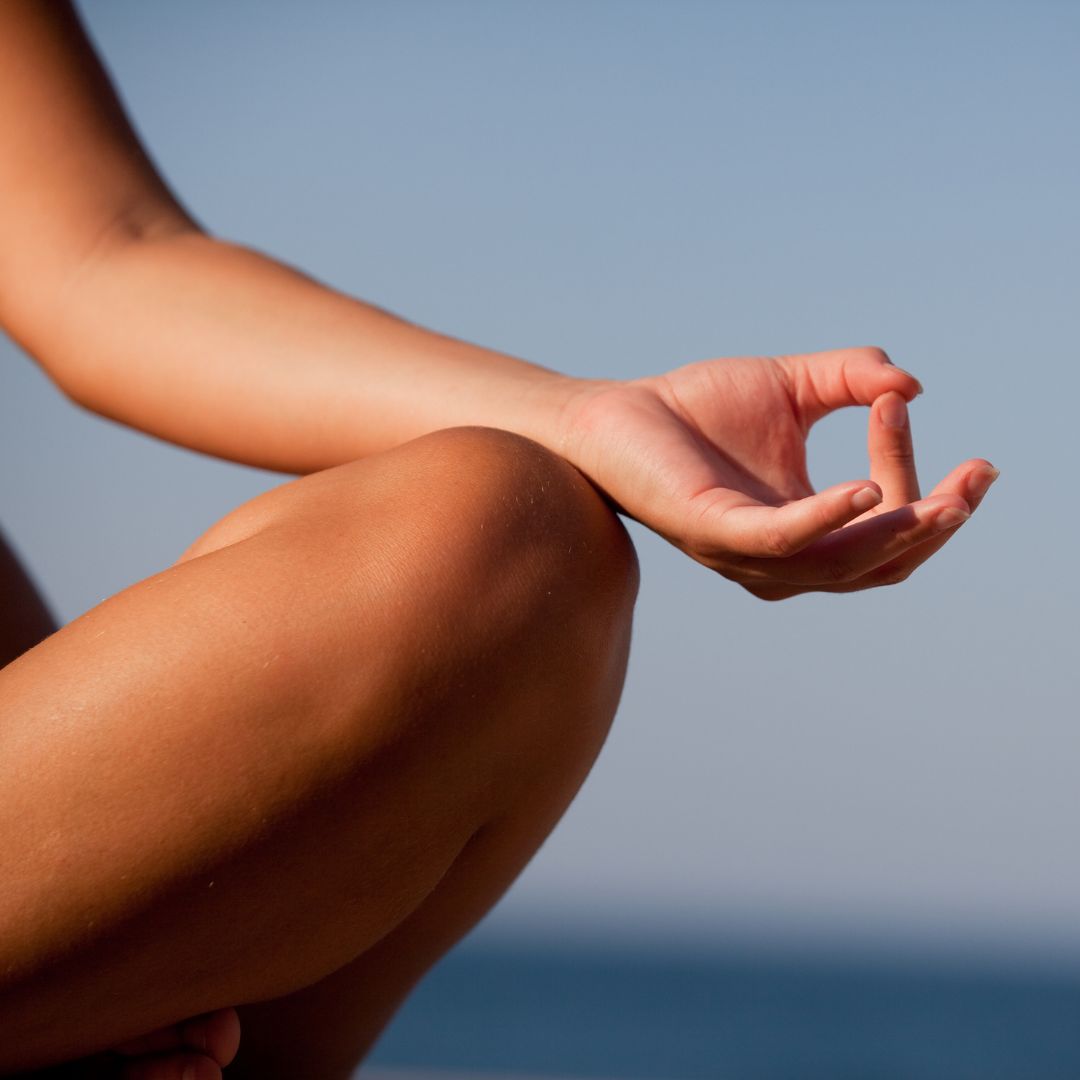 A person sitting cross-legged outdoors with their hand resting on their knee in a meditation gesture, against a blurred blue sky background.