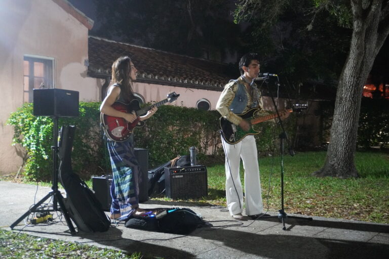 Two musicians performing outdoors at night, one playing the guitar and the other singing into a microphone.