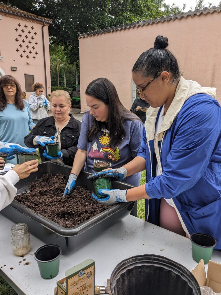 Group of people engaging in a gardening workshop, filling pots with soil from a large tub outdoors as part of the programs for students at Vizcaya.