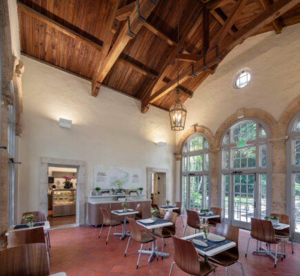 The dining area at Vizcaya Cafe boasts wooden ceiling beams and large arched windows. It features table seating surrounded by potted plants, with a doorway leading to a counter where a variety of items are beautifully displayed, enhancing the shop's charm.