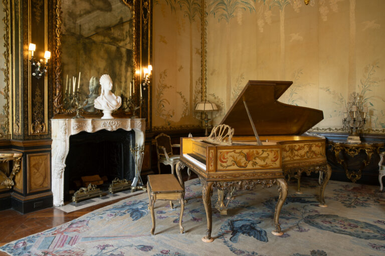 A lavish room with ornate décor, featuring a grand piano, a white bust above a fireplace, elegant wall designs, and vintage furniture on a patterned rug.