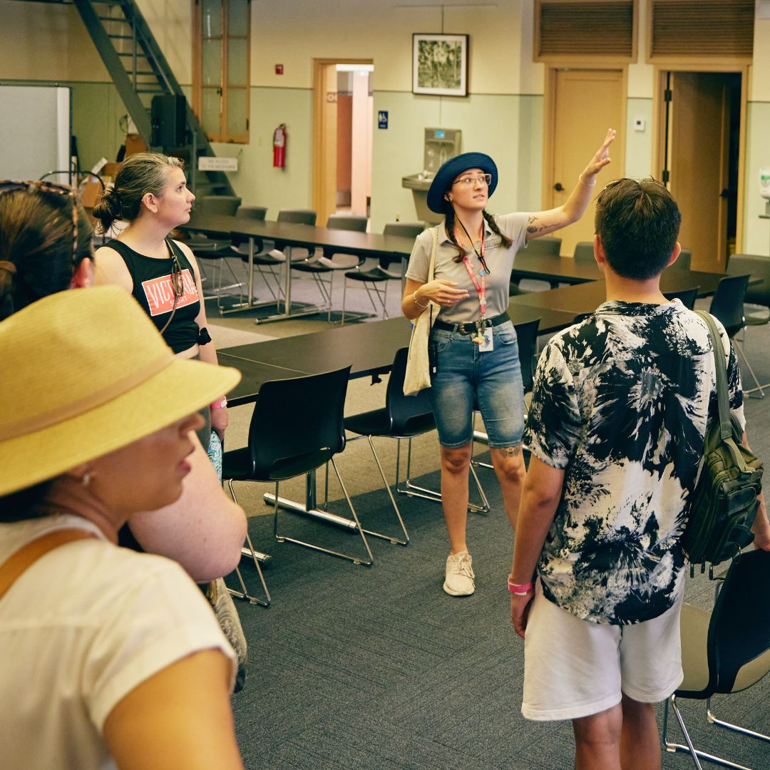 A woman wearing a hat and lanyard speaks to a small group in a classroom or conference room with empty chairs and tables.