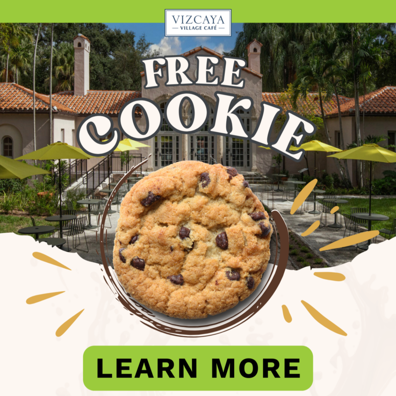 A chocolate chip cookie is centered over an outdoor cafe setting. The text reads "FREE COOKIE" with "LEARN MORE" below, inviting you to explore new stories and programs at Vizcaya Village history events.