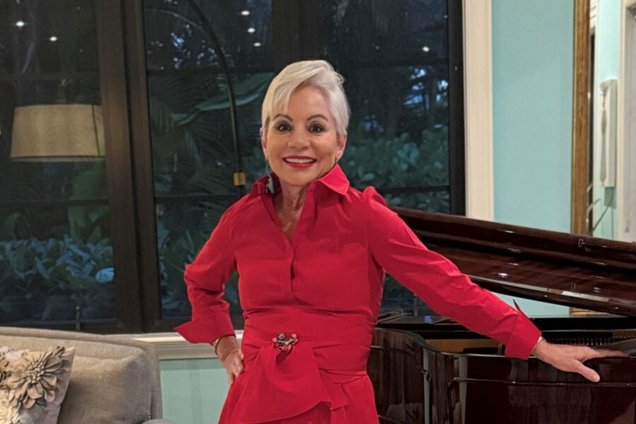 An older woman with short gray hair, wearing a bright red outfit, stands smiling with one hand resting on a grand piano in a living room.