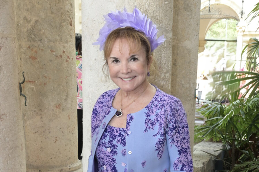 Swanee DiMare wearing a lavender floral dress and matching feathered headpiece stands indoors near stone pillars and greenery, smiling at the camera.