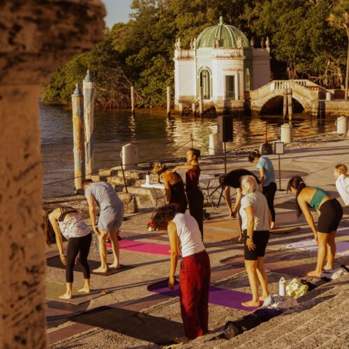 A group of people practice yoga on mats outdoors near a waterfront, with a historic pavilion and trees in the background.