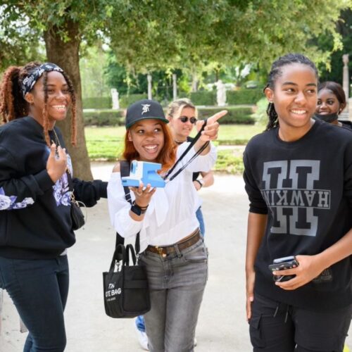 A group of young people smiling and laughing outdoors, with one person holding a camera and pointing, surrounded by trees in a park setting.