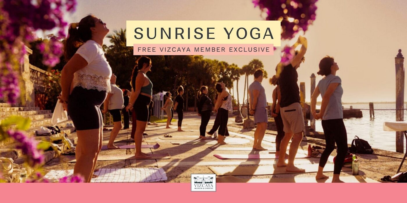 A group of people practice yoga outdoors on mats near the water at sunrise, with trees, flowers, and columns in the background.