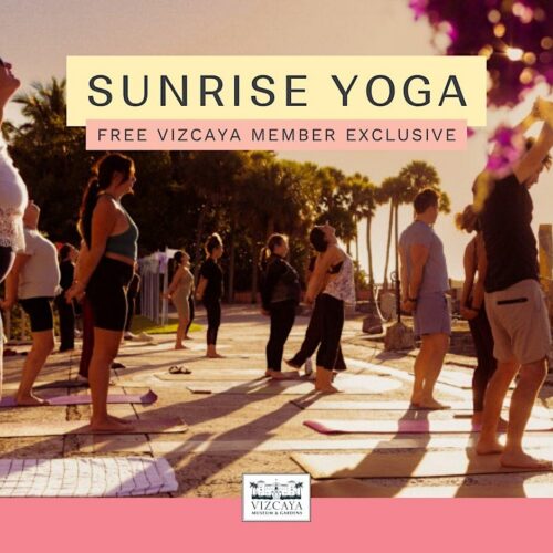 A group of people practice yoga outdoors on mats near the water at sunrise, with trees, flowers, and columns in the background.