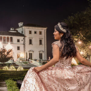 Woman in a sparkling ball gown and tiara stands outside a grand, illuminated mansion at night, holding her dress and smiling.