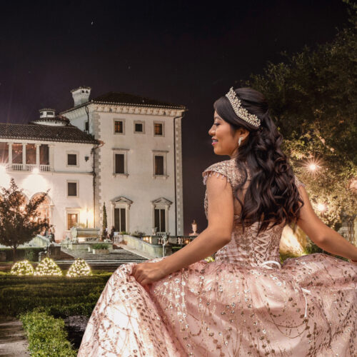 Woman in a sparkling ball gown and tiara stands outside a grand, illuminated mansion at night, holding her dress and smiling.