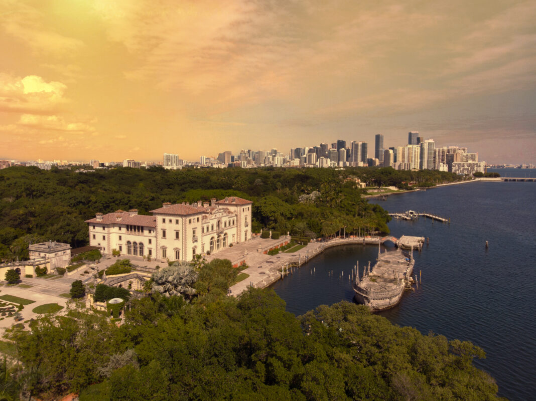Aerial view of a large historic mansion by the water, surrounded by greenery, with a city skyline in the background under an orange sky.