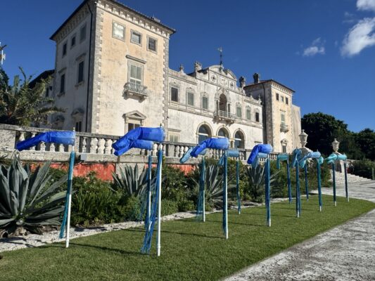 A historic stone mansion with arched windows stands behind a row of blue fabric art installations set on poles in a landscaped garden.