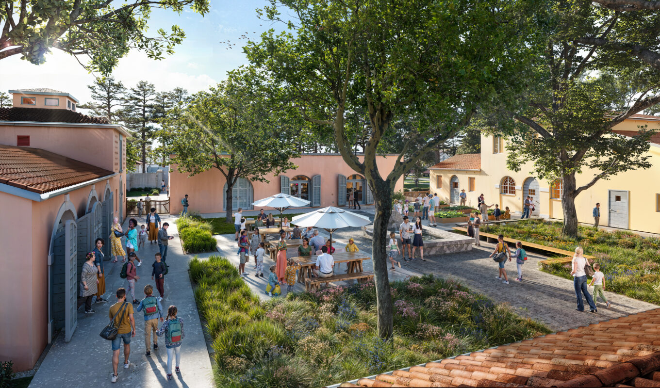 People gather in a sunny courtyard with trees, benches, and tables with umbrellas between pastel-colored buildings. Some are walking, while others sit and talk.