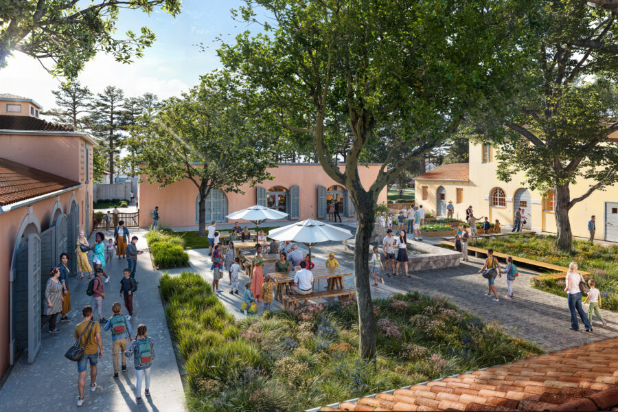 People gather in a sunny courtyard with trees, benches, and tables with umbrellas between pastel-colored buildings. Some are walking, while others sit and talk.