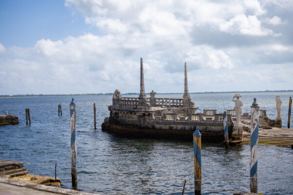 Stone barge with decorative sculptures and columns in the water near a shoreline, under a partly cloudy sky—an elegant backdrop for weddings and events at Vizcaya.