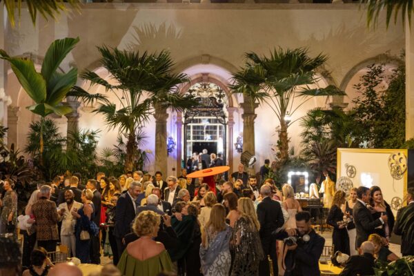 A large group of people in formal attire gather outdoors at night for a social event, surrounded by palm trees and the elegant architecture of Vizcaya—making it a stunning setting for weddings and events at Vizcaya.
