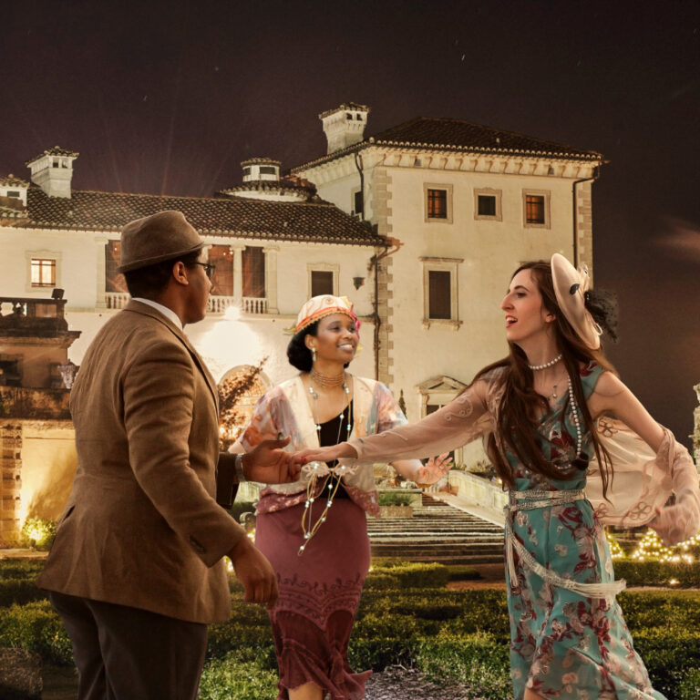 Three people in 1920s-style clothing stand and interact in front of a lit-up historic building at night.