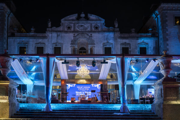 An outdoor event setup with a chandelier, tables, and stage in front of an illuminated historic building at night—perfect for unforgettable weddings and events at Vizcaya.
