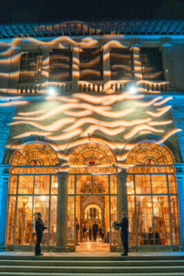A grand building with arched glass doors is illuminated by warm lights inside and swirling patterns outside at night. Two people stand at the entrance, evoking the magical ambiance of weddings and events at Vizcaya.