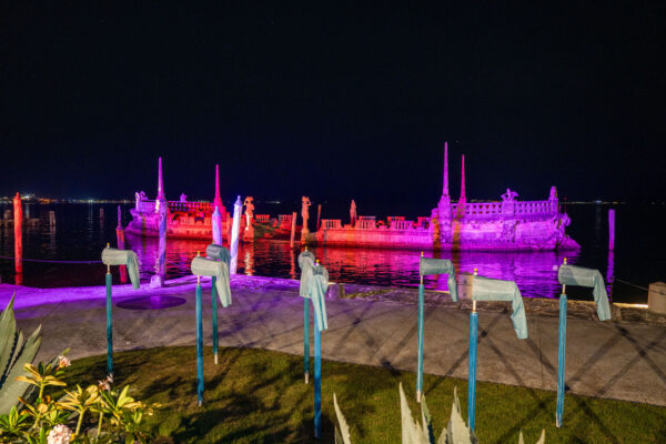 A stone barge structure at Vizcaya glows with pink and purple lights at night, setting a magical scene for weddings and events, with several blue-painted objects lined up in the foreground.