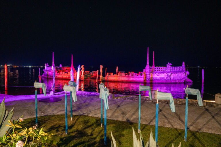 A stone barge structure at Vizcaya glows with pink and purple lights at night, setting a magical scene for weddings and events, with several blue-painted objects lined up in the foreground.