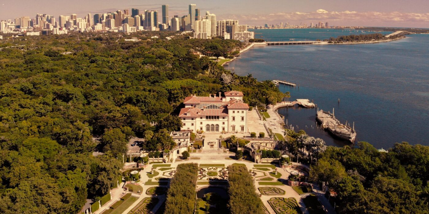 Aerial view of Vizcaya Museum and Gardens by Biscayne Bay, with Miami skyline and high-rise buildings in the background.