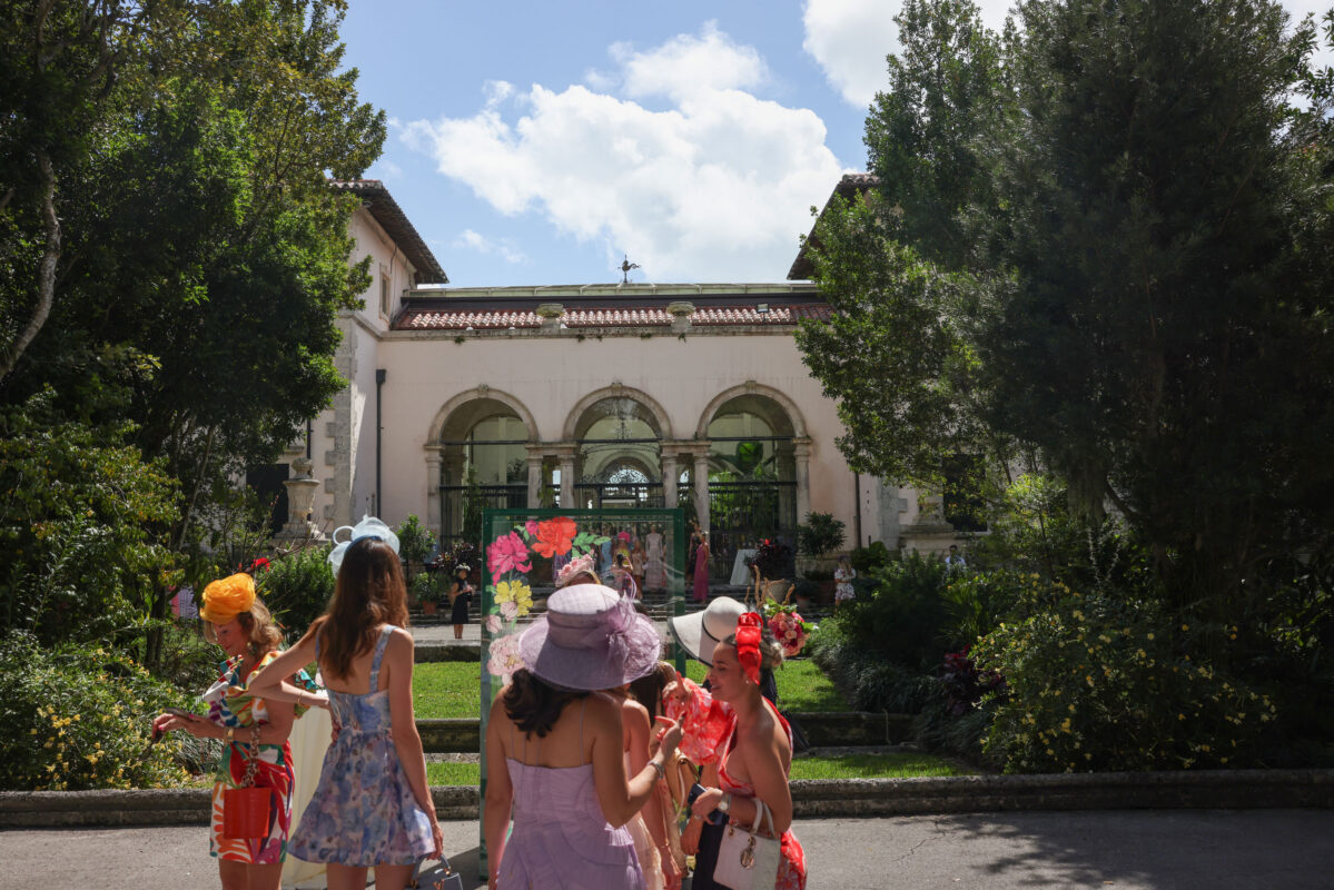 A group of women in colorful dresses and hats stand chatting in front of a historic building with arched windows and a lush garden.