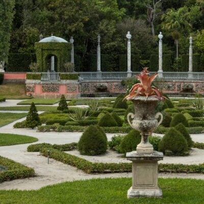 A formal garden with manicured hedges, a stone urn with a red bromeliad, a gazebo covered in greenery, and a columned pergola in the background.
