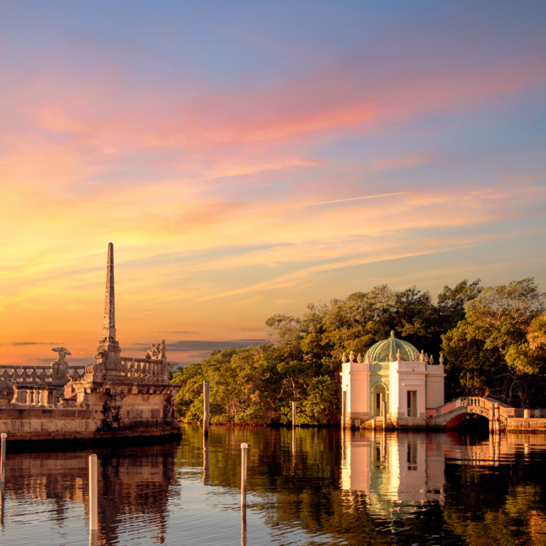 A historic waterside pavilion and stone barge at sunset, with calm water reflecting the sky and surrounding greenery.