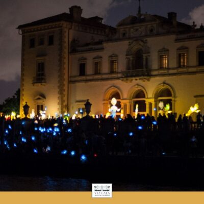 A large crowd holds blue lights at night in front of a lit historic building with arched windows and a balcony.