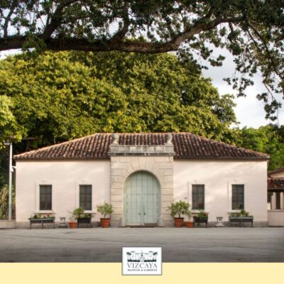 A small, single-story building with a tiled roof and arched mint-green door, surrounded by potted plants and large trees, at Vizcaya Museum & Gardens.