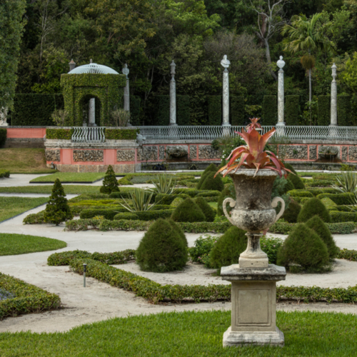 Formal garden with neatly trimmed hedges, topiary bushes, a stone urn planter in the foreground, and a gazebo with columns in the background.