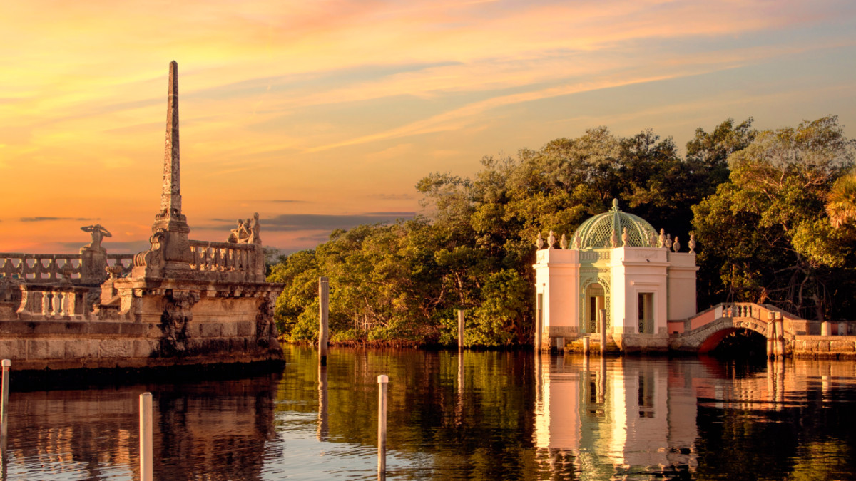 A sunset view of a waterside pavilion with a green dome and a stone structure featuring sculptures, surrounded by trees and reflecting on calm water.