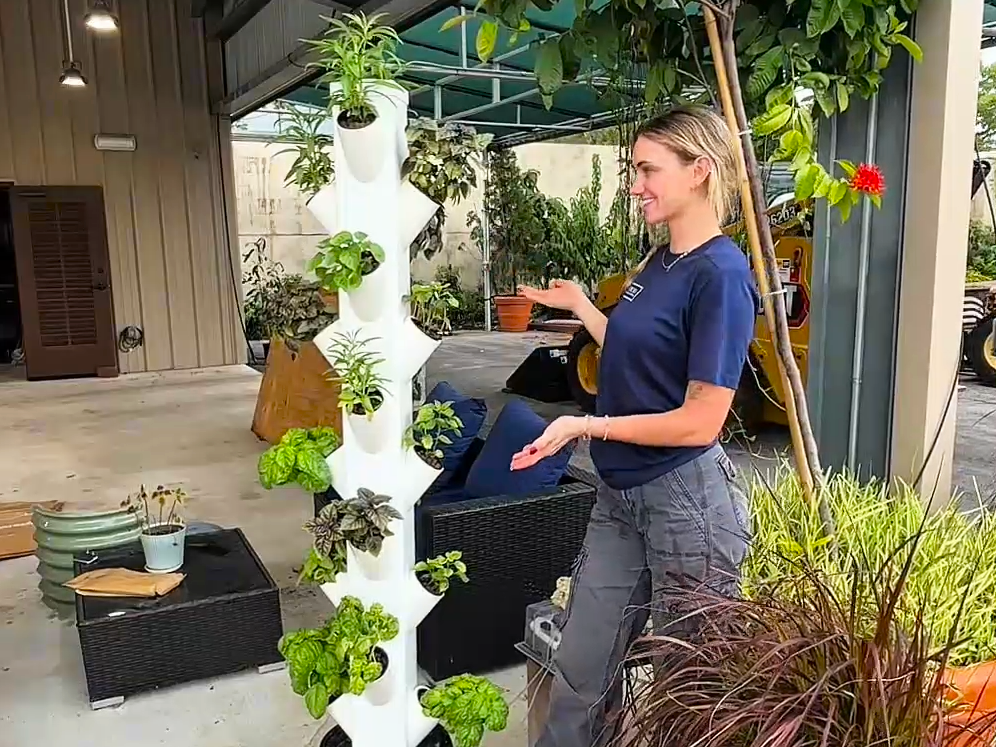 A person stands beside a vertical white planter with various herbs and plants, outdoors under a covered area with garden supplies and potted plants nearby.