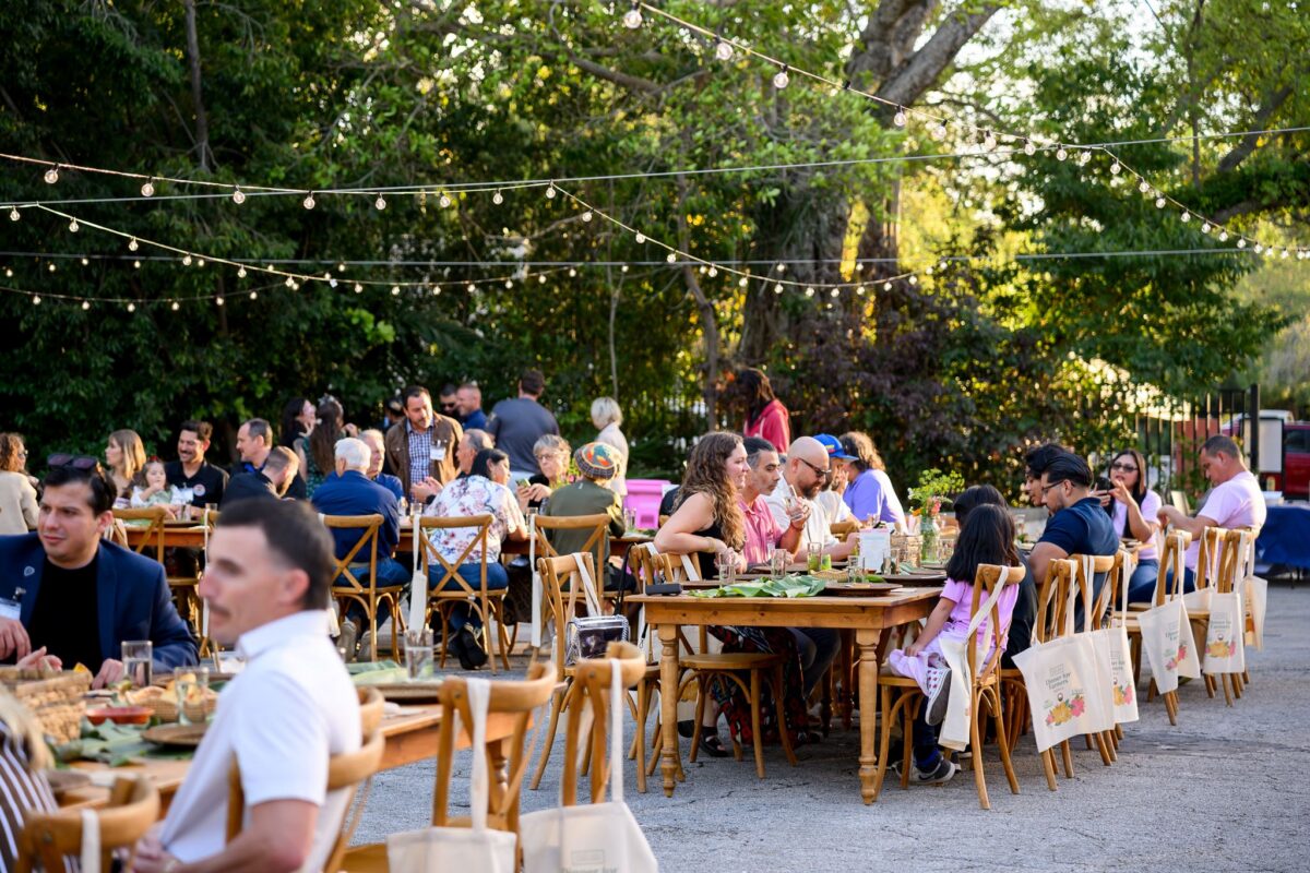People sit at wooden tables outdoors under string lights, eating and talking in a relaxed garden setting with greenery in the background.
